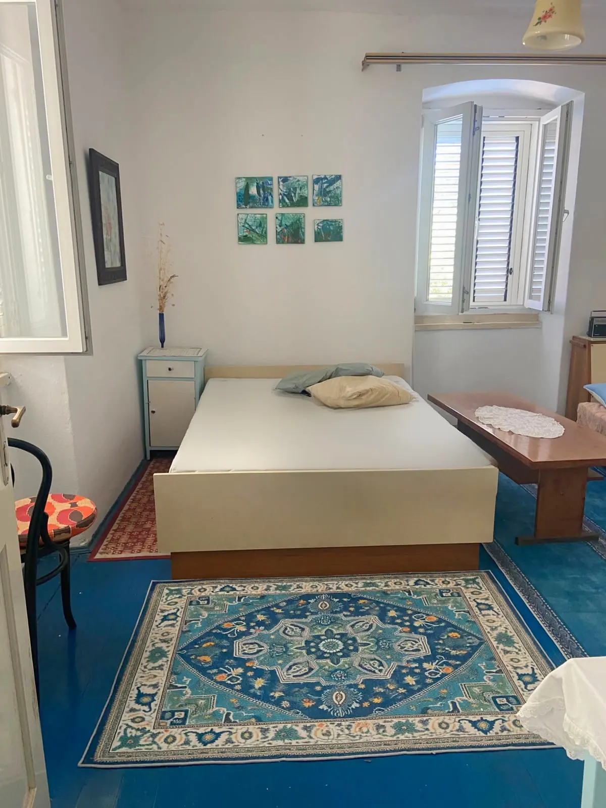Bedroom with a blue wooden floor, a patterned rug, a double bed and green paintings by the window