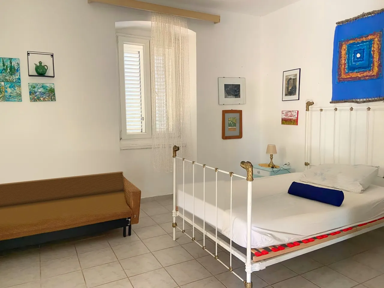 Bright room with a white iron bed, a striped sofa, a window with a lace curtain and paintings on the walls