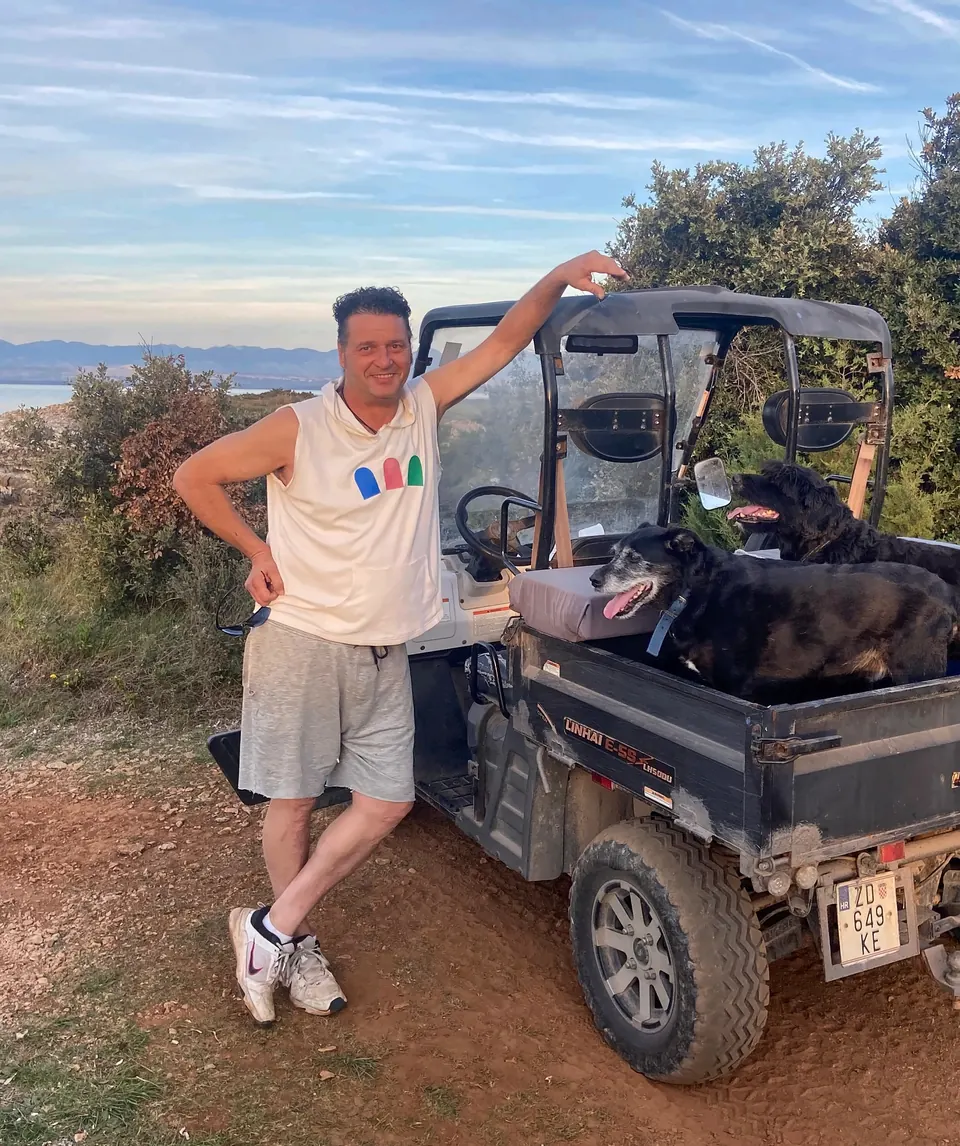 Denis Kleva leaning on his electric utility cart with two dogs in the back, above the sea on Olib