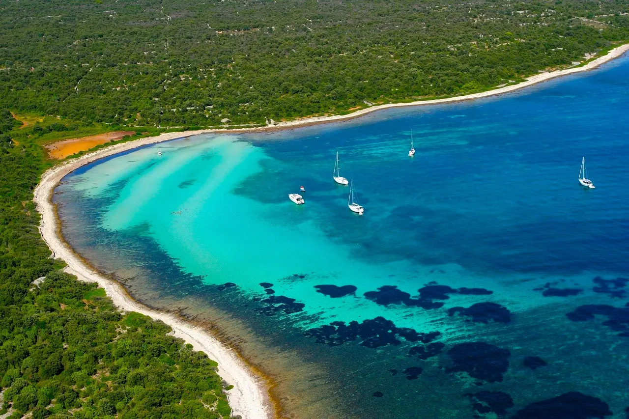 A long sandy bay with turquoise, shallow water and anchored sailboats