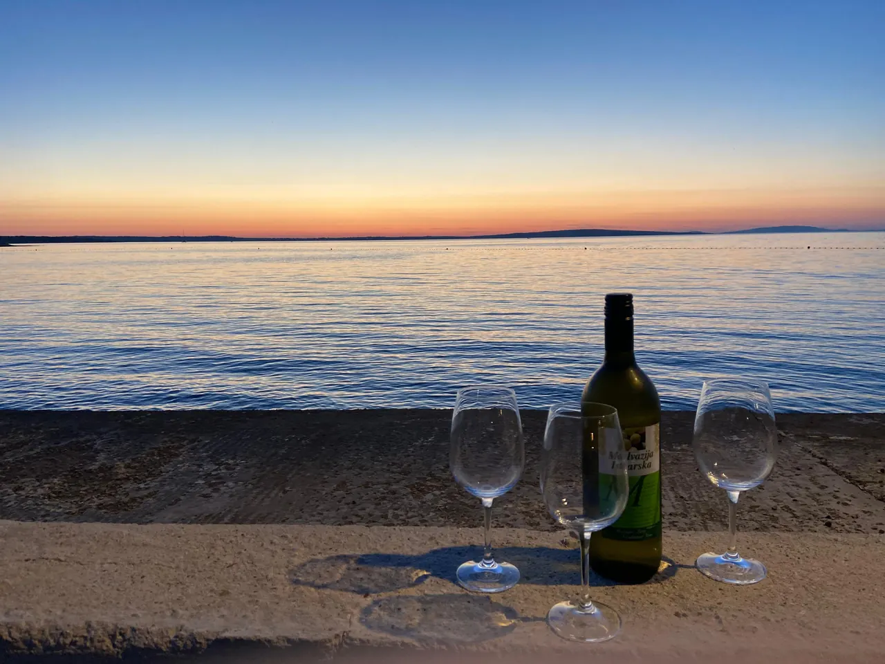 Bottle of local wine and three glasses on a stone wall by the sea at sunset